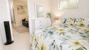 Ein Schlafzimmer mit Bett, Kommode, Spiegel, Nachttisch, Lampe und einem Fenster mit Jalousien.