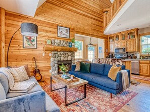 A cozy cabin interior with a stone fireplace, wooden walls, a blue sofa, a dining area, and a kitchen with wooden cabinets. - NEW! Mt. Hood Escape | Pool, Bunk Room & Dogs Okay (Rhododendron)