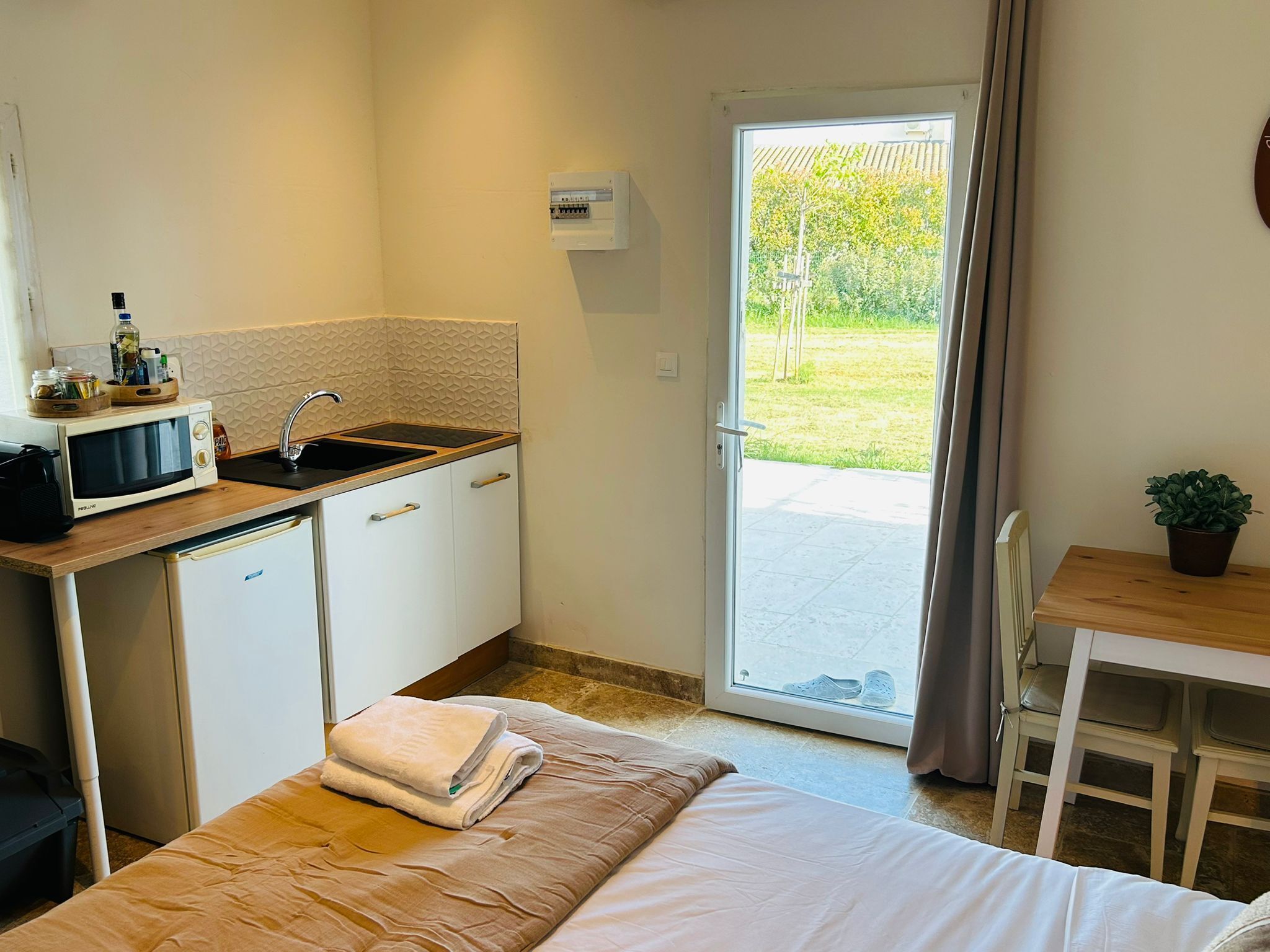 A compact kitchen with a microwave, sink, and cabinets, a bed with a folded towel, and a view of a garden through a sliding door.