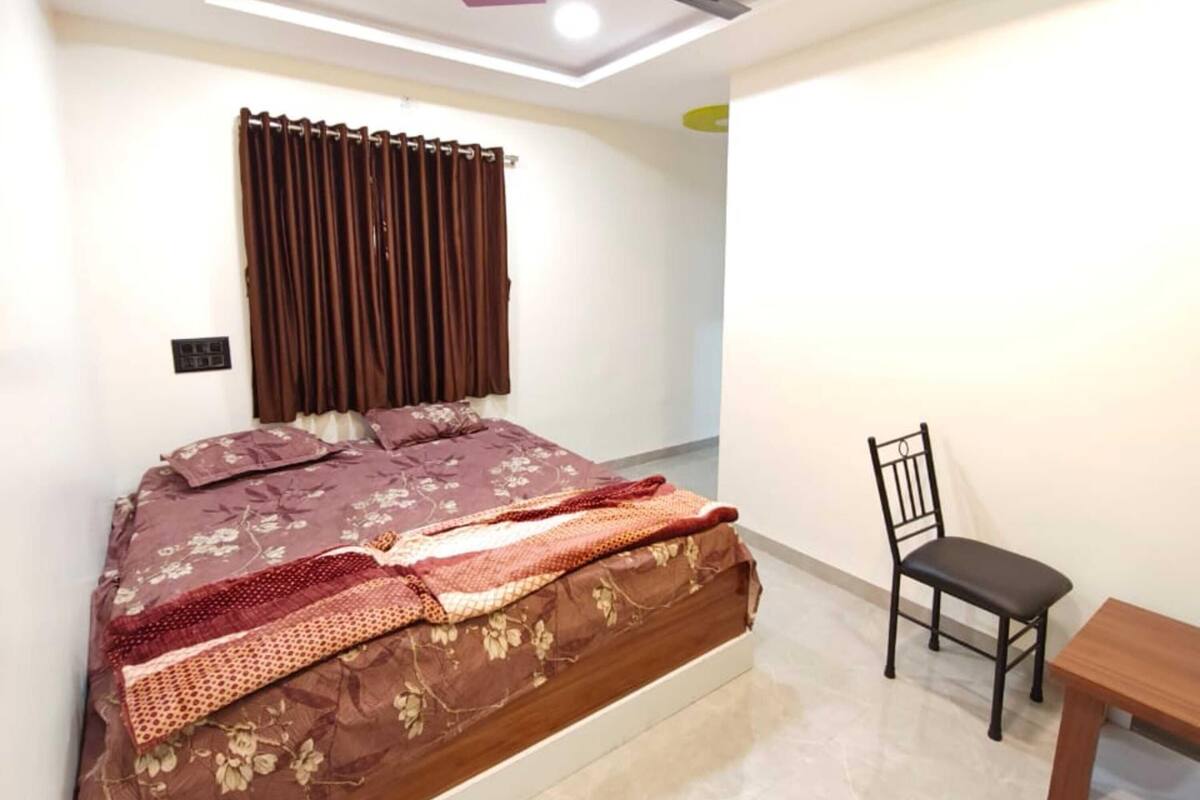 A bedroom with a bed, a chair, a table, a ceiling fan, and a light fixture.