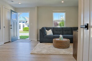 Ein modernes Wohnzimmer mit einem grauen Sofa, einem Korbtisch und Blick auf ein Haus durch eine Tür.