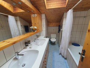 Een badkamer met een houten plafond, een grote spiegel, twee wasbakken, een toilet en een bad.