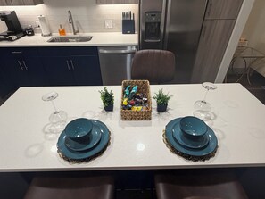 A modern kitchen with a white countertop, stainless steel appliances, and a dining area with teal plates and wine glasses. - Stylish One-Bedroom Getaway Near Everything (Addison)