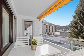 Un balcon avec une table et des chaises blanches, un panier en osier rempli de fleurs et une vue sur des immeubles et des arbres couverts de neige.