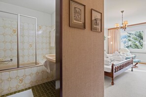 Une salle de bain avec une baignoire, des toilettes et une douche. On y trouve un lit avec une tête de lit en bois et une fenêtre avec des rideaux.