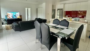 Una sala de estar moderna con un sofá negro, una mesa de comedor de vidrio con sillas, un televisor y una zona de cocina con armarios blancos y detalles en rojo.