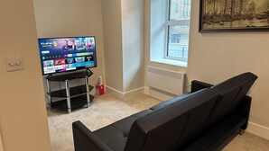 Una sala de estar con un televisor sobre un mueble bajo, un sofá negro, una ventana con persianas y un radiador.