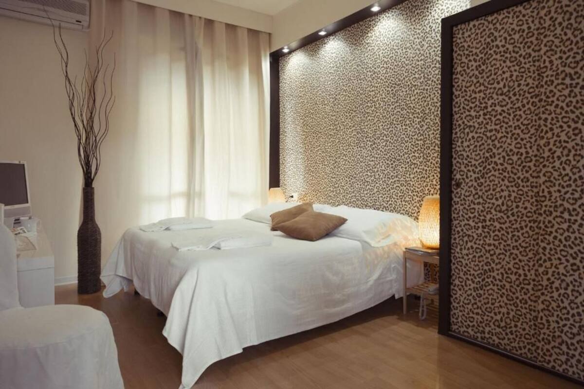 A hotel room with a bed, bedside tables, a lamp, a vase with branches, and a window with curtains.