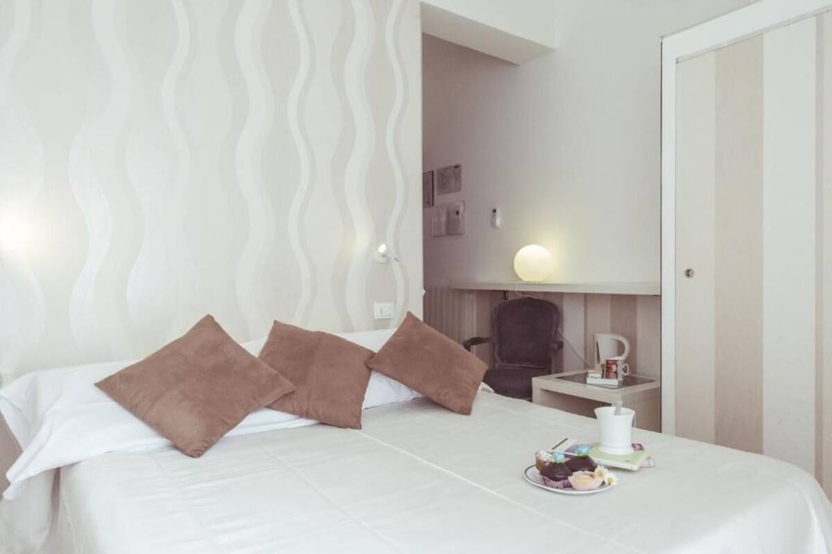A hotel room with a bed, pillows, a bedside table with a lamp, a chair, and a small table with a cup and saucer.