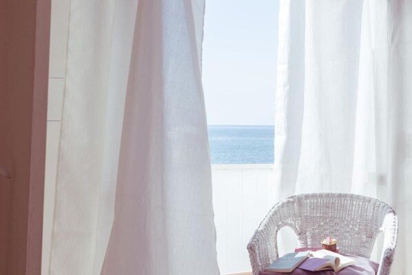 A room with a view of the sea, a wicker chair, and a table with books and a cupcake.