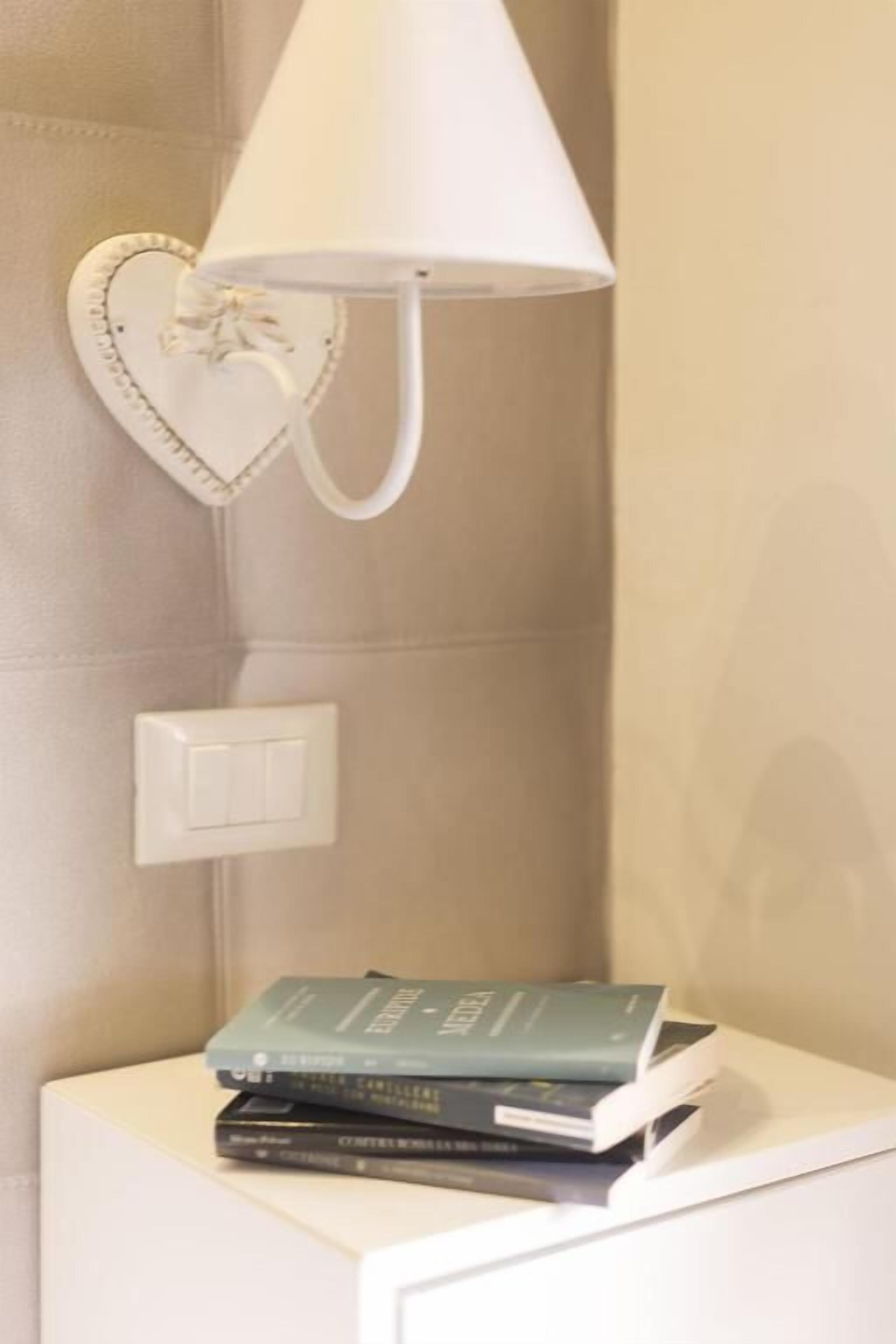 Un comodino con una lampada e una decorazione a forma di cuore, una pila di libri e un interruttore a muro.