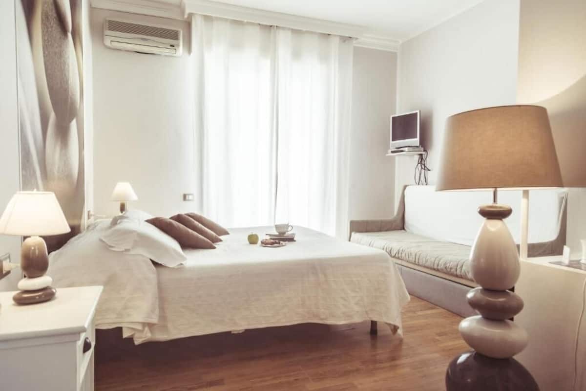 A hotel room with a bed, bedside tables, a TV, a lamp, and a window with curtains.