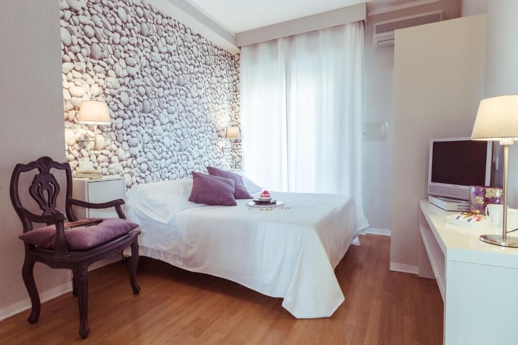 Un dormitorio con pared de piedra, cama con ropa blanca, una silla de madera, mesita de noche con lámpara y televisor sobre un mueble.