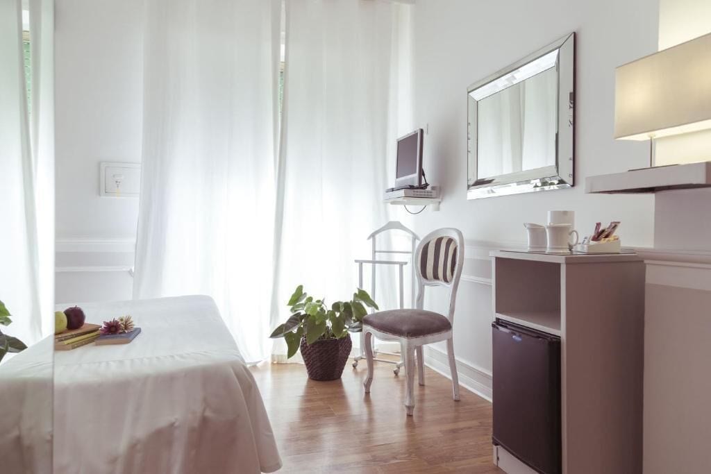 A hotel room with a bed, a desk, a chair, a TV, a mirror, a refrigerator, and a potted plant.