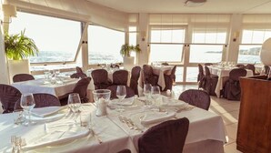 Un comedor con mesas dispuestas para una comida, ventanas amplias con vista al mar y plantas en macetas.