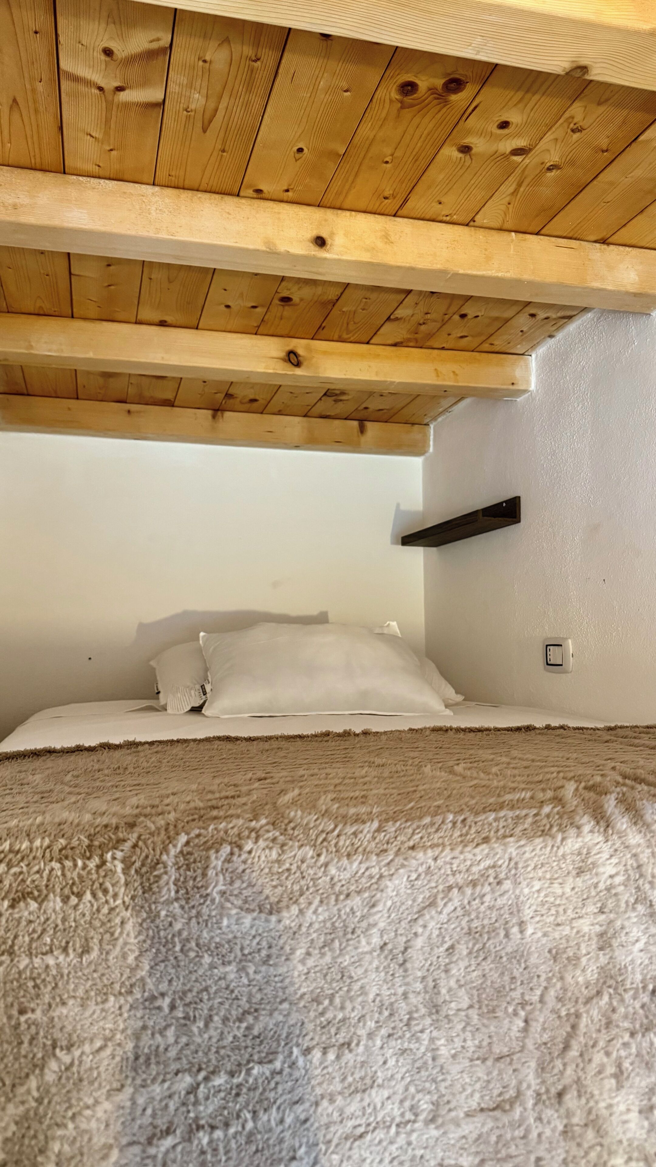 A room with a wooden ceiling, a bed with white bedding, a fluffy rug, and a wall-mounted electrical outlet.