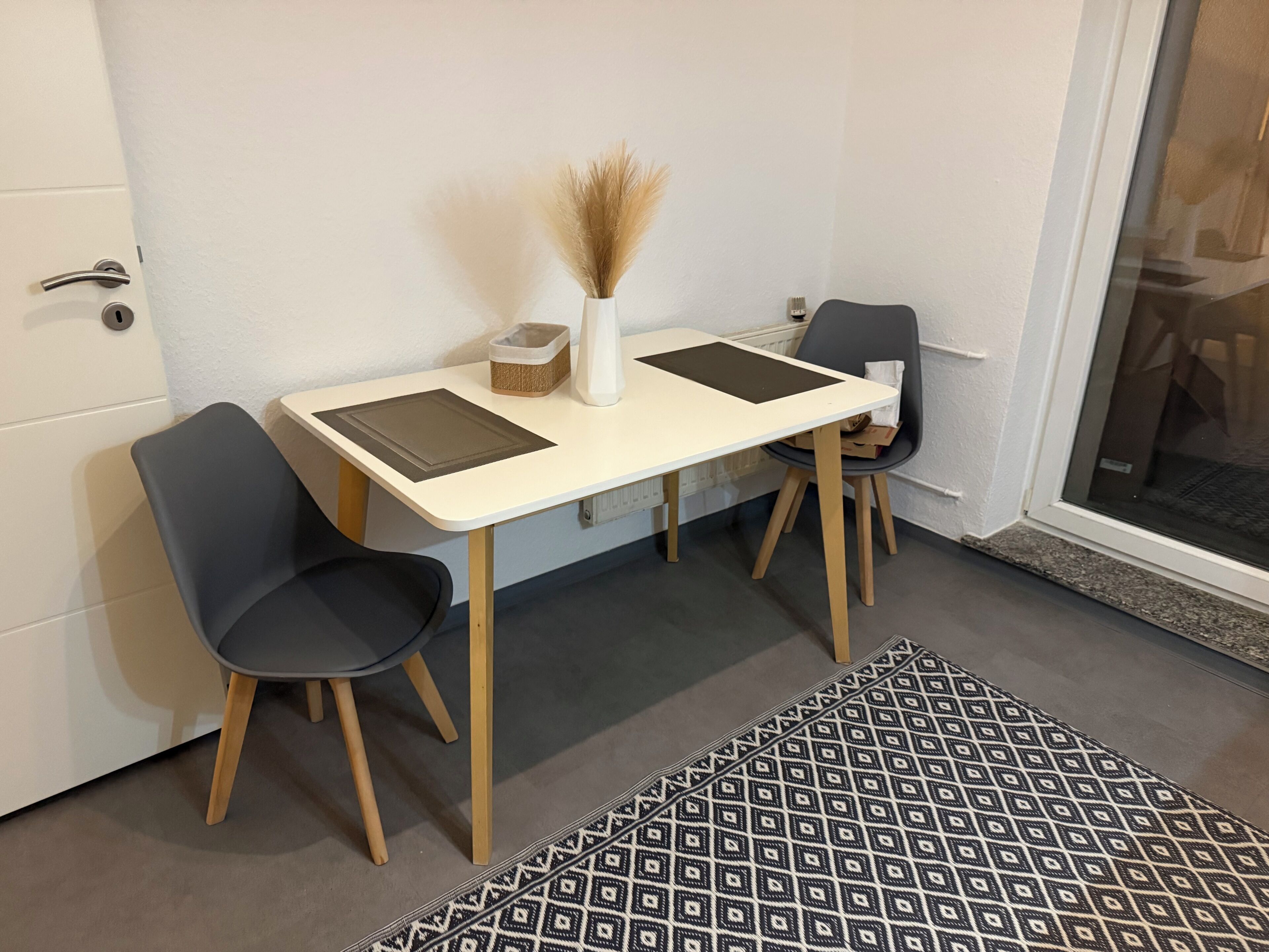 Ein kleiner, moderner Raum mit einem weißen Tisch, zwei Stühlen, einer Vase mit Trockenpflanzen, einem gemusterten Teppich und einer Tür mit Schlüsselloch.