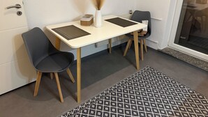 Ett litet, modernt rum med ett vitt bord, två stolar, en vas med torkade växter, en mönstrad matta och en dörr med ett nyckelhål.