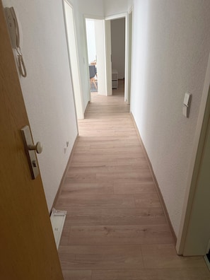 Ein Korridor mit Holzdielen, weißen Wänden und einer Tür links.