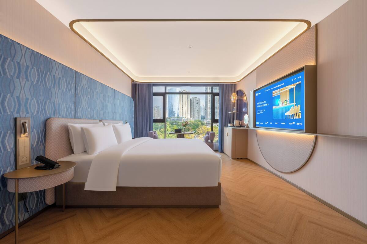 A modern hotel room with a large bed, a television, and a view of the cityscape.