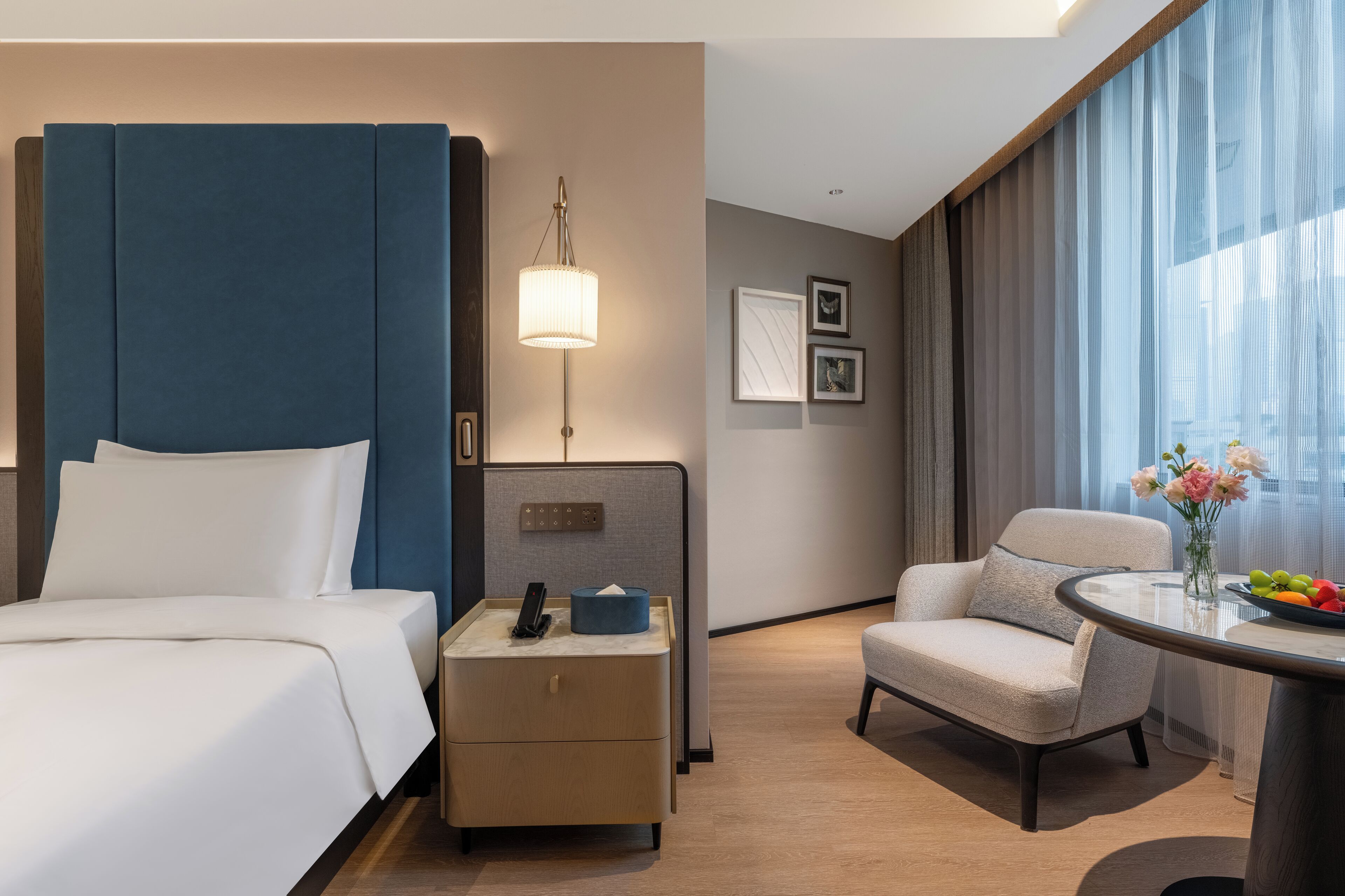 Habitación de hotel con una cama grande, una mesita de noche con lámpara, una silla, una pequeña mesa con flores y una ventana con cortinas.