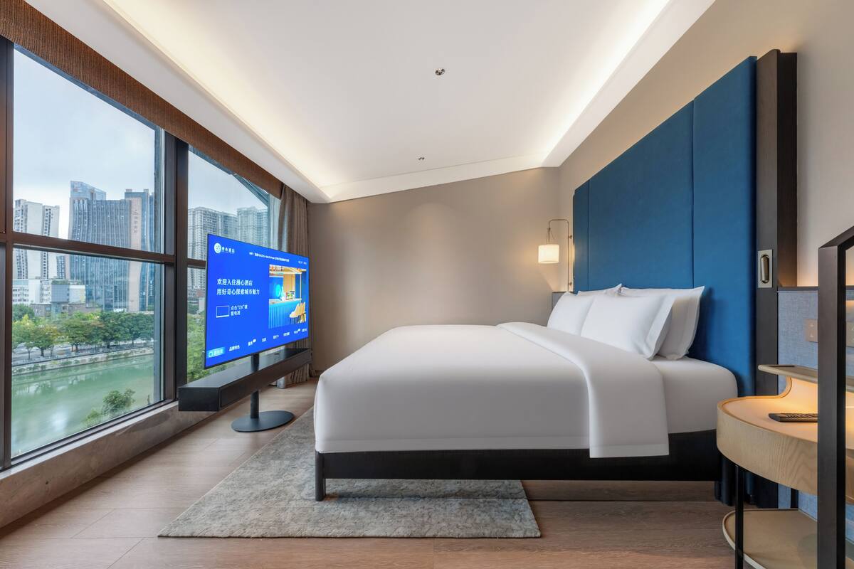 A modern hotel room with a large window, a bed, a TV, and a desk.