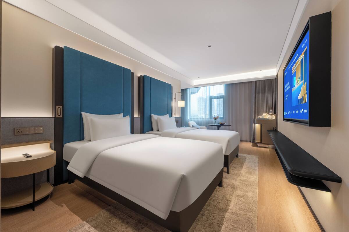 A hotel room with two beds, a TV, a desk, and a window with a city view.