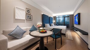 Una habitación de hotel moderna con sofá, cama, mesa de comedor con fruta, televisor y vista a la ciudad.