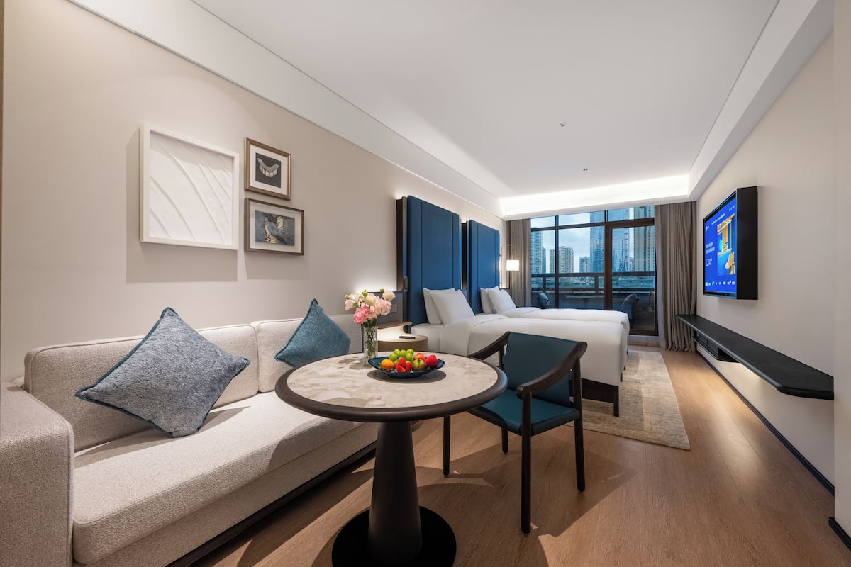 A modern hotel room with a sofa, a bed, a dining table with fruit, a TV, and a city view.