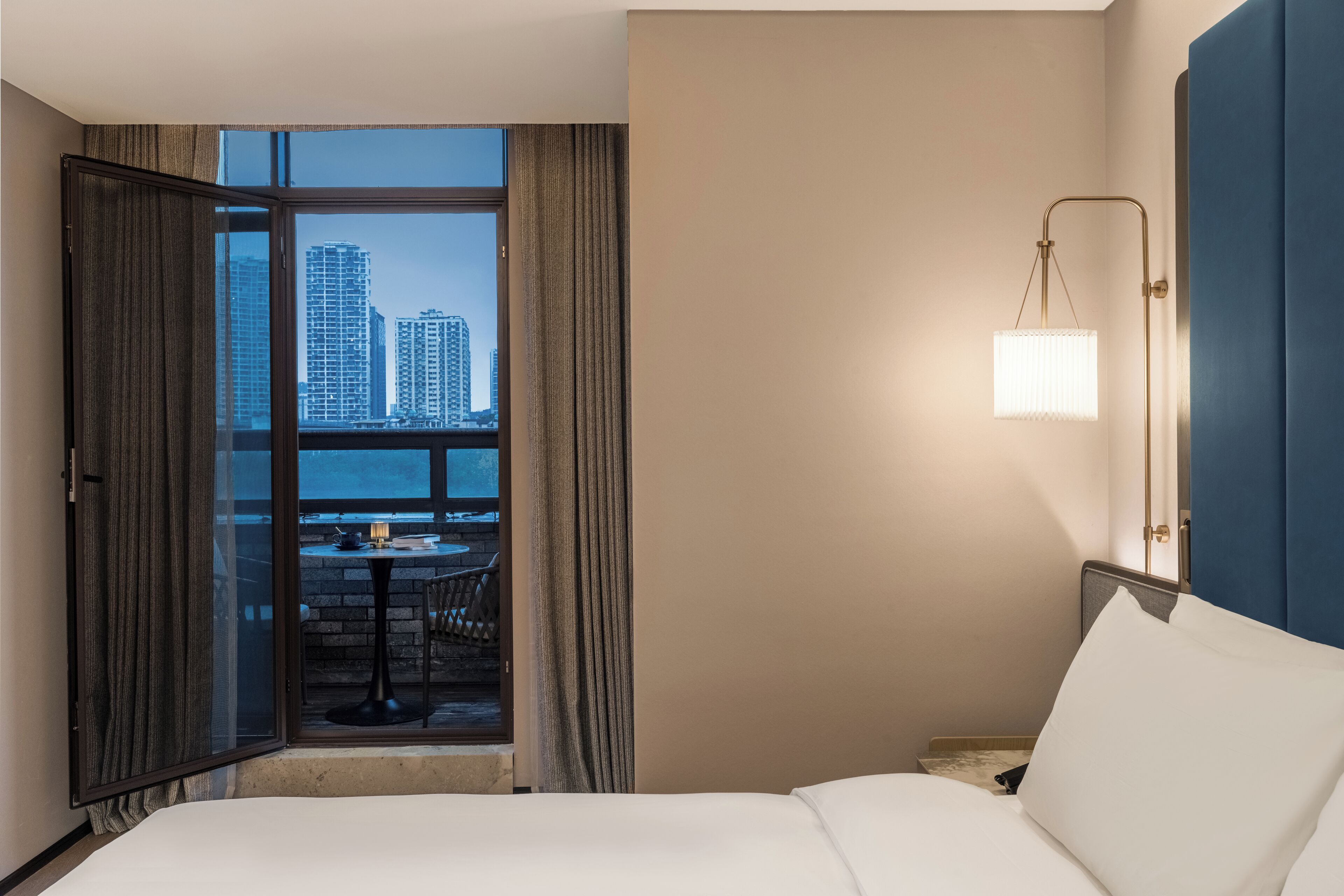 Un cuarto de hotel con cama, mesita de noche, lámpara, vista de edificios altos a través de una ventana y balcón con mesa y sillas.