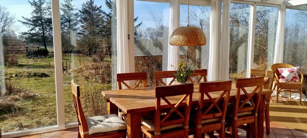 Un hivernacle amb taula i cadires de fusta, una làmpada de fibra i vista a una zona verda amb arbres.