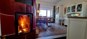 Een kamer met een open haard, houten vloeren, een zithoek met stoelen en een tafel, en een keuken met keukenkastjes.