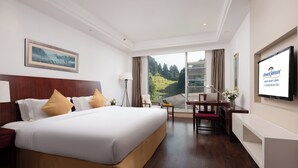 A hotel room with a large bed, a desk, a chair, a TV, and a window with a view of greenery. - Howard Johnson Lushan Jiahao (Jiujiang)