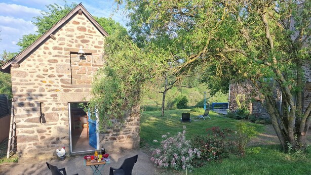A stone building with a door and windows, a small table with chairs, and a garden with grass and plants.