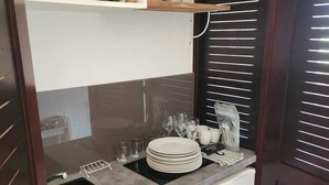 Een keukentje met een magnetron, een kleine koelkast, een spoelbak en een aanrecht met gestapelde borden en glazen.