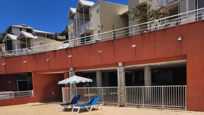 Een zwembad met helder water, omgeven door een rood gebouw met balkons en een overdekt terras met ligbedden.