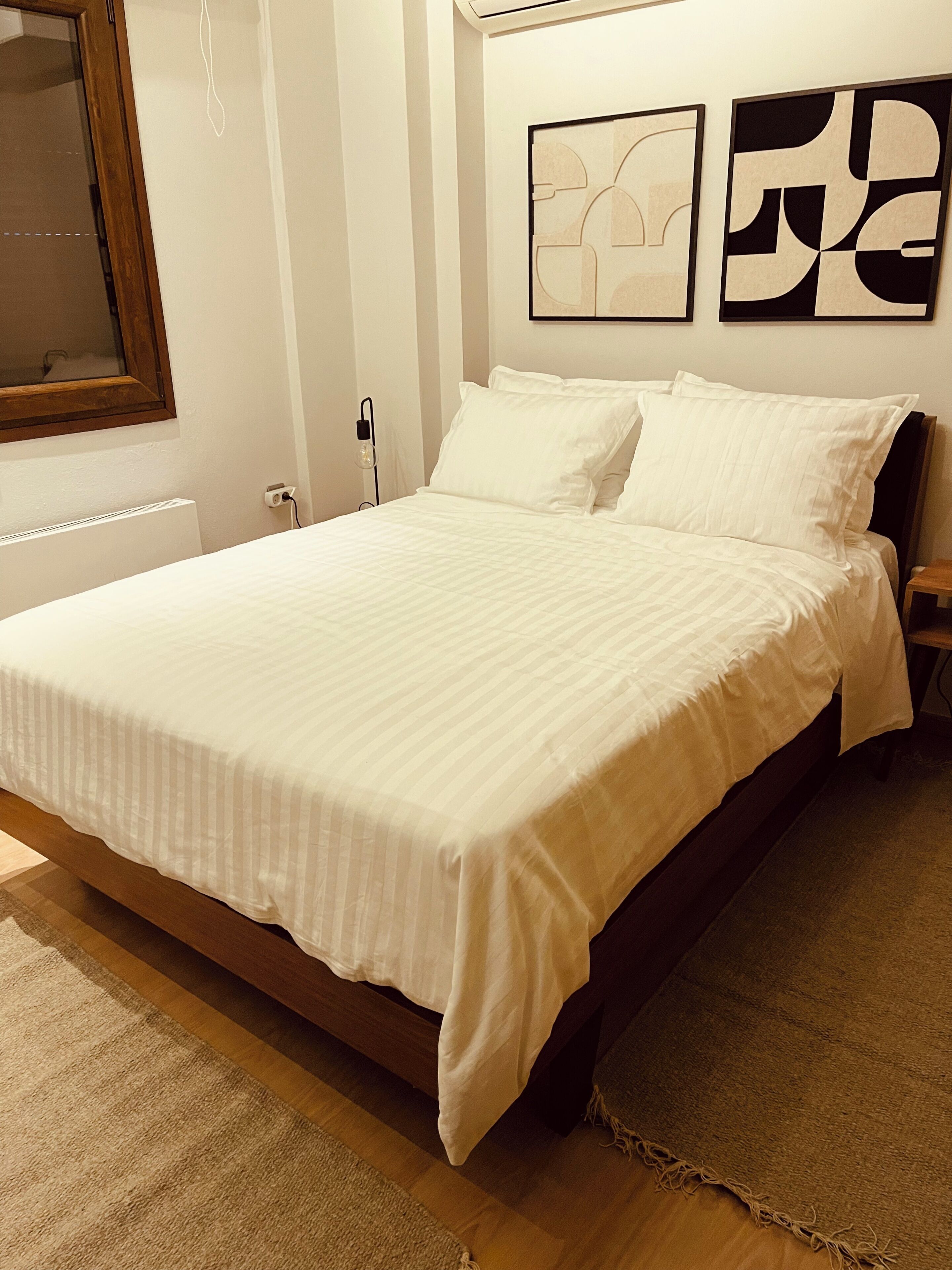 Cama bem arrumada com roupa de cama branca, cabeceira de madeira e uma pequena mesa de cabeceira.