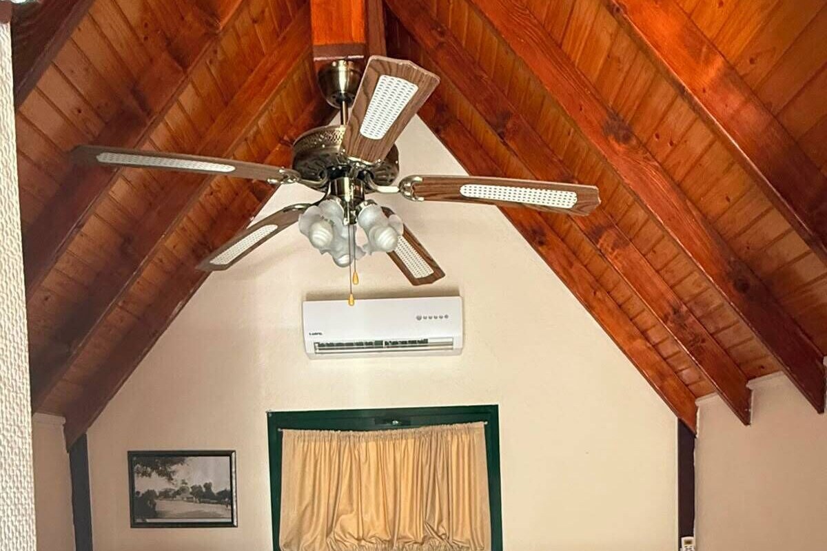 Un dormitorio con cama, ventilador de techo, equipo de aire acondicionado, mesita de noche con lámpara y un cuadro colgado en la pared.