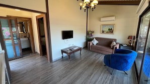 A room with a wooden floor, a brown sofa, a blue chair, a TV, and a chandelier. - Seal beach (Mordoğan)