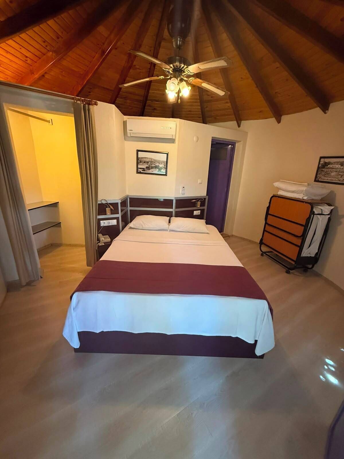 Un dormitorio con cama, ventilador de techo, equipo de aire acondicionado, una puerta y una pequeña mesa con una silla.