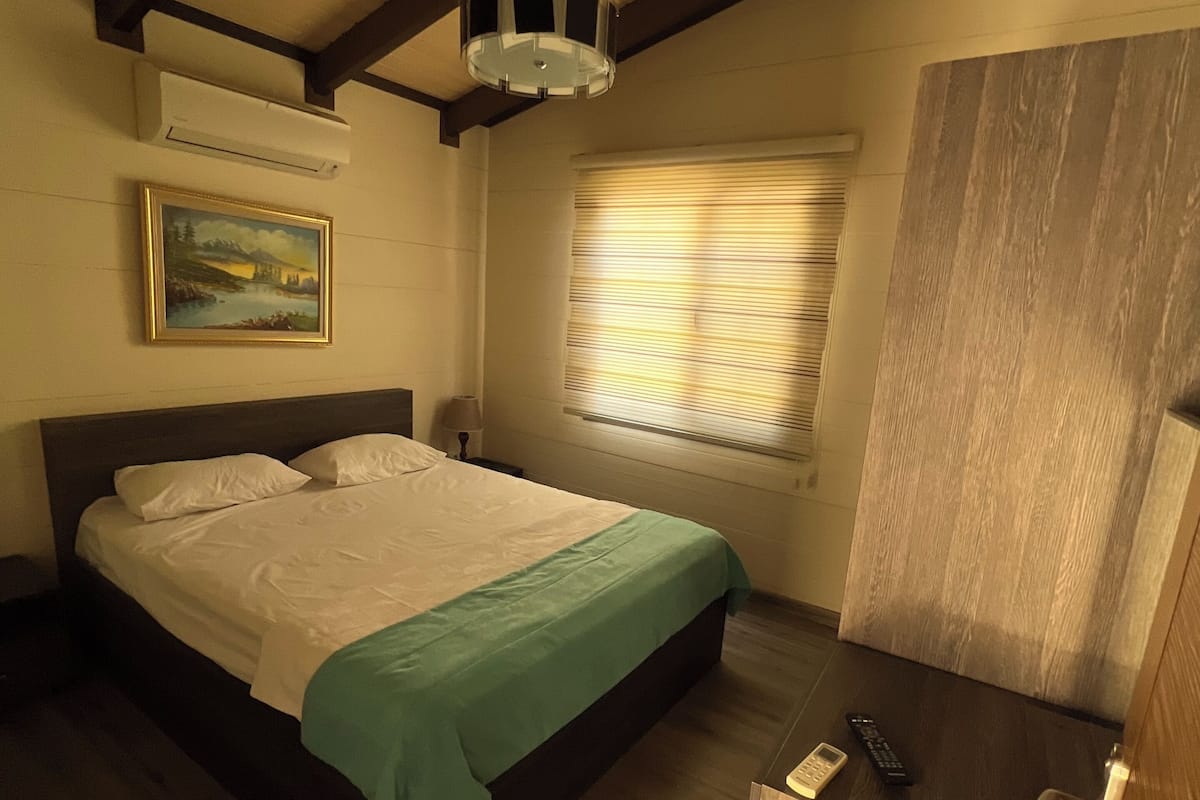 Une chambre Ă coucher avec un lit, un tableau, une fenĂȘtre avec des stores, une armoire en bois et une table de chevet avec une tĂ©lĂ©commande.