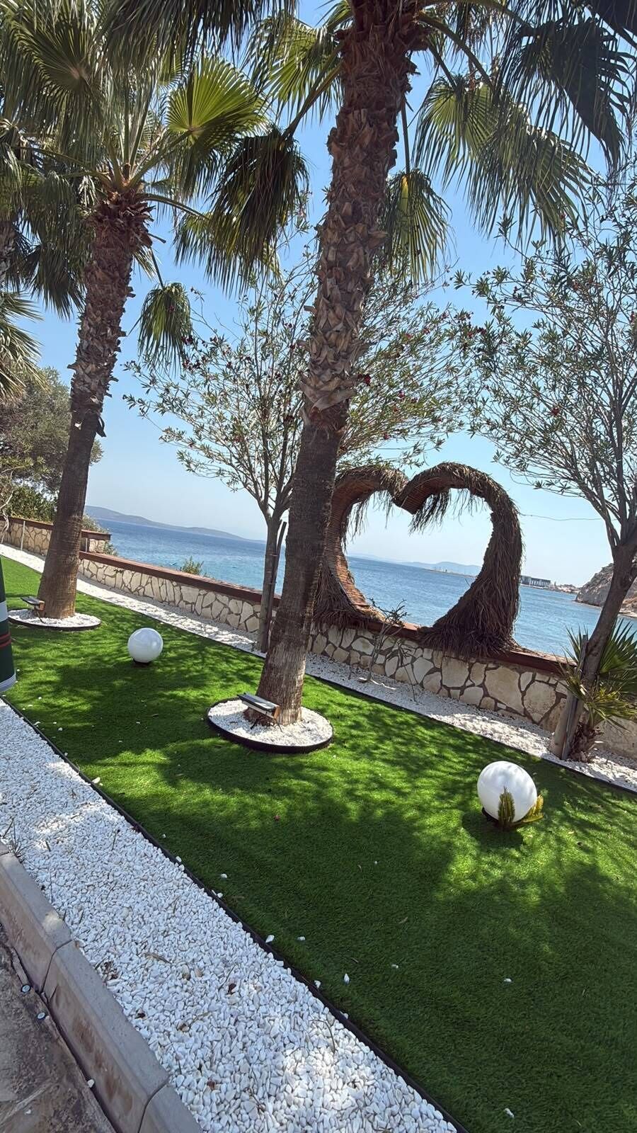 Zona ajardinada con palmeras, un arreglo de ramas con forma de corazón y vista al mar.