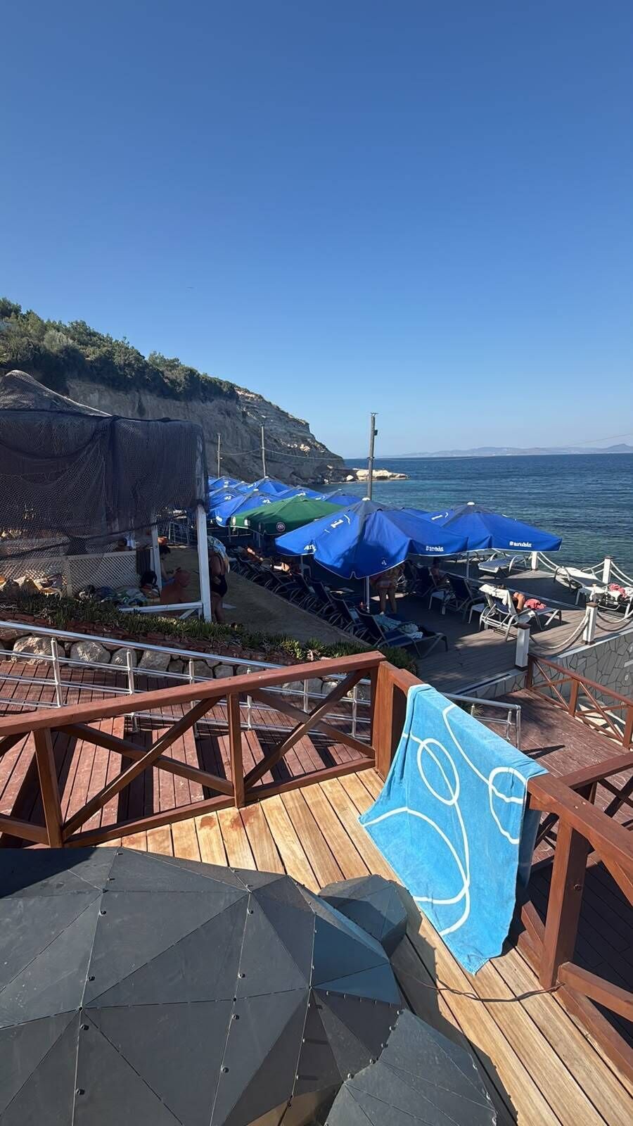 A wooden deck with outdoor seating and umbrellas, overlooking a coastal area with a clear sky.