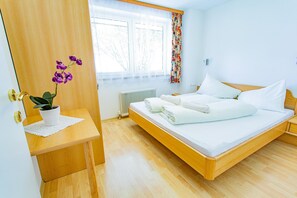 Ein Schlafzimmer mit einem hölzernen Bett, einem Nachttisch mit einer Topfpflanze, einem Fenster mit durchsichtigen Vorhängen und einer Heizung.