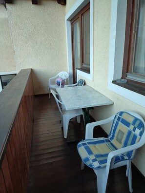 Een balkon met een houten vloer, een witte tafel, twee witte plastic stoelen en een raam met houten kader.