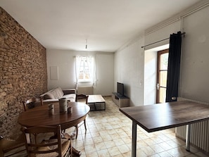A room with a stone wall, a round wooden table with chairs, a sofa, a TV, and a window with curtains.