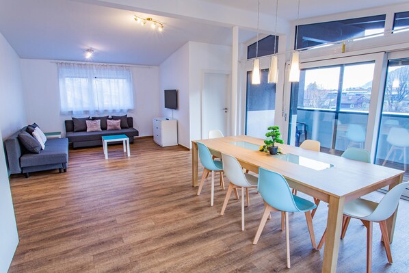 Ein modernes Wohnzimmer mit einer Couch, einem Esstisch mit Stühlen, einem Fernseher und einem Fenster mit Vorhängen.