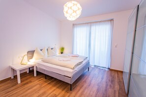Ein Schlafzimmer mit Bett, Nachttisch, Lampe und einem großen Fenster mit Vorhängen.