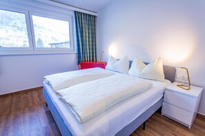 Ein Doppelbett mit weißer Bettwäsche, ein Nachttisch mit Lampe und ein Fenster mit Vorhängen.