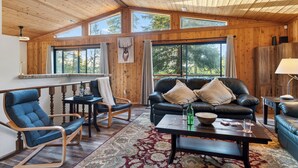 Un salón espacioso con paredes y techo de madera, ventanas amplias, un sofá de cuero negro, un sillón azul y una mesa de centro de madera.